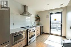 Kitchen featuring wall chimney range hood, stainless steel appliances, light stone counters, open shelves, and light wood-style floors -