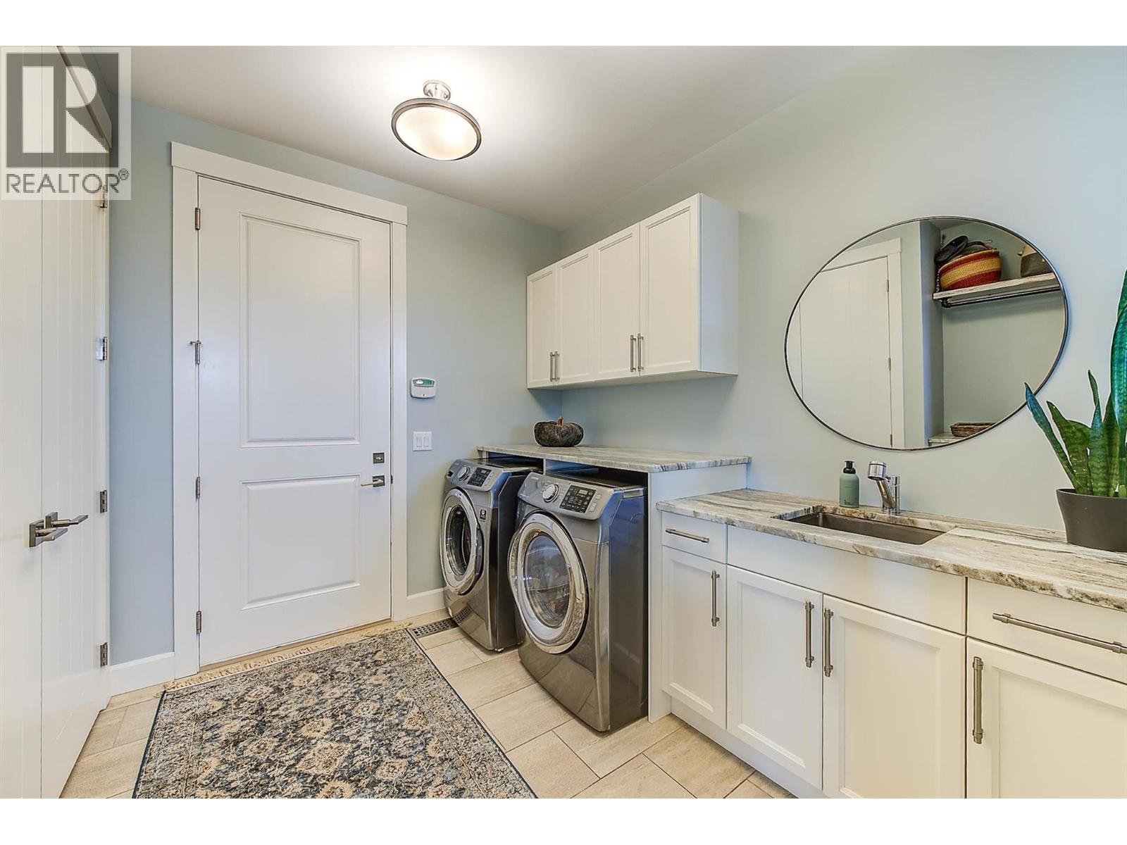 The laundry/mud room features white cabinetry with granite countertops, a deep stainless steel sink, large-format tile flooring, and front-loading stainless steel Samsung steam washer and dryer. - 738 Highpointe Drive, Kelowna, BC - Indoor Photo Showing Laundry Room