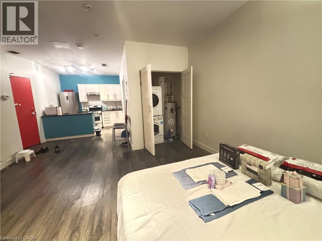 Bedroom with freestanding refrigerator, stacked washing machine and dryer, dark wood-style floors, water heater, and lofted ceiling - 25 Main Street Unit# 302, Cambridge, ON - Indoor Photo Showing Other Room