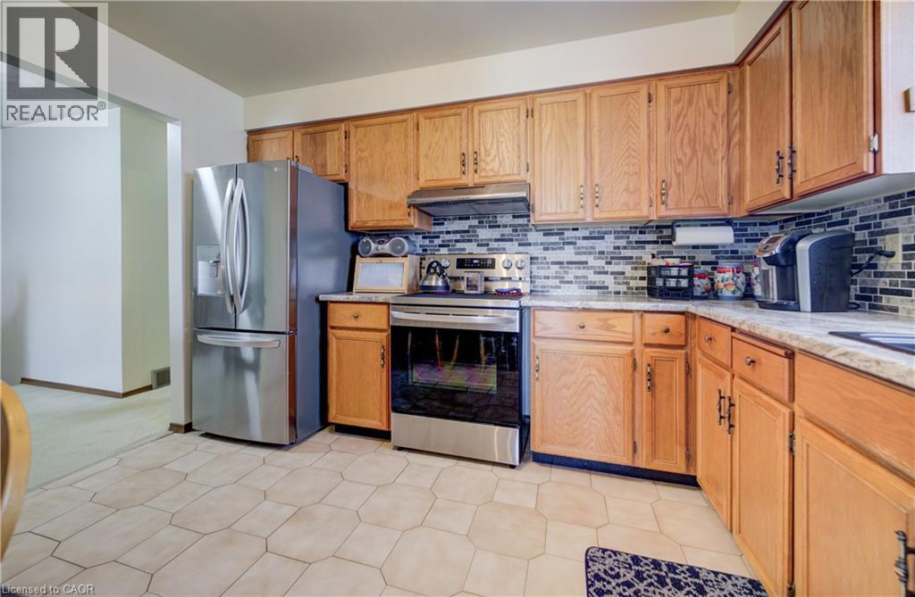 Kitchen featuring stainless steel appliances, under cabinet range hood, brown cabinetry, light stone counters, and backsplash - 42 Ruffian Road, Brantford, ON - Indoor Photo Showing Kitchen