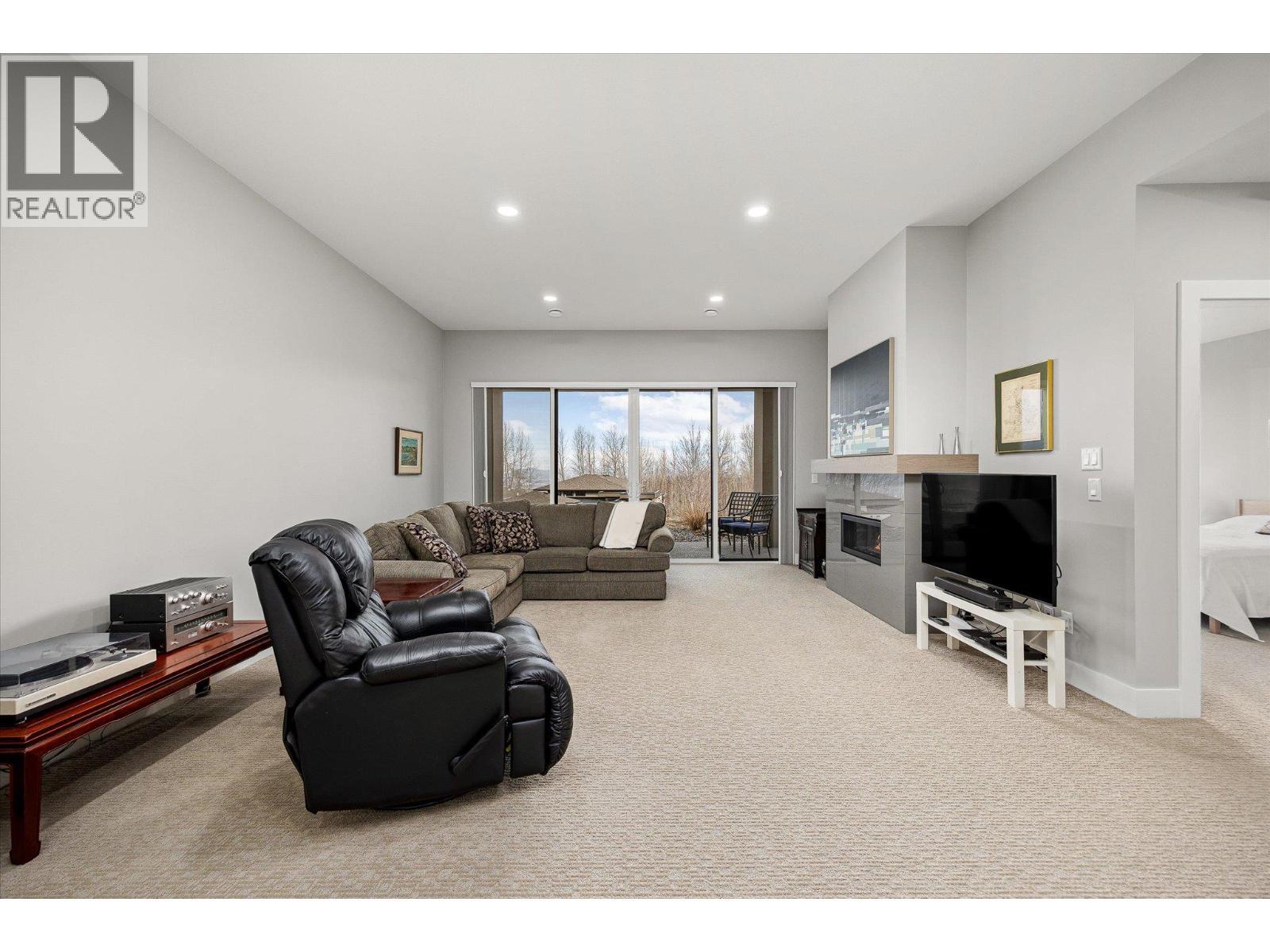 Lake views from the lower level! Spacious recreation space with tall ceilings featuring a gas fireplace with tile surround all opening to a covered patio - 468 Sparrow Hawk Court, Kelowna, BC - Indoor Photo Showing Living Room