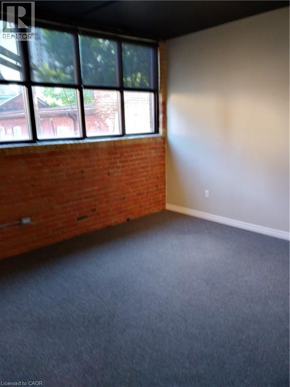Empty room featuring brick wall and dark carpet - 12 Ferguson Avenue N, Hamilton, ON