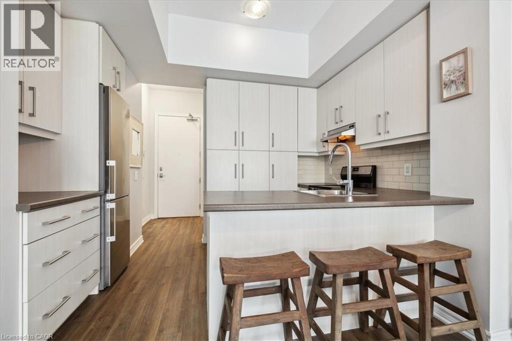Kitchen with a kitchen bar, a peninsula, dark countertops, decorative backsplash, and dark wood-style floors - 40 Esplanade Lane Unit# 102, Grimsby, ON - Indoor Photo Showing Kitchen
