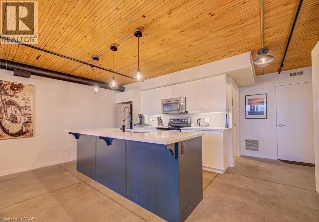 Dual tone kitchen with decorative light fixtures, a kitchen island with sink, stainless steel microwave, wooden ceiling, and black range with electric cooktop - 85 Spruce Street Unit# 103, Cambridge, ON - Indoor Photo Showing Kitchen