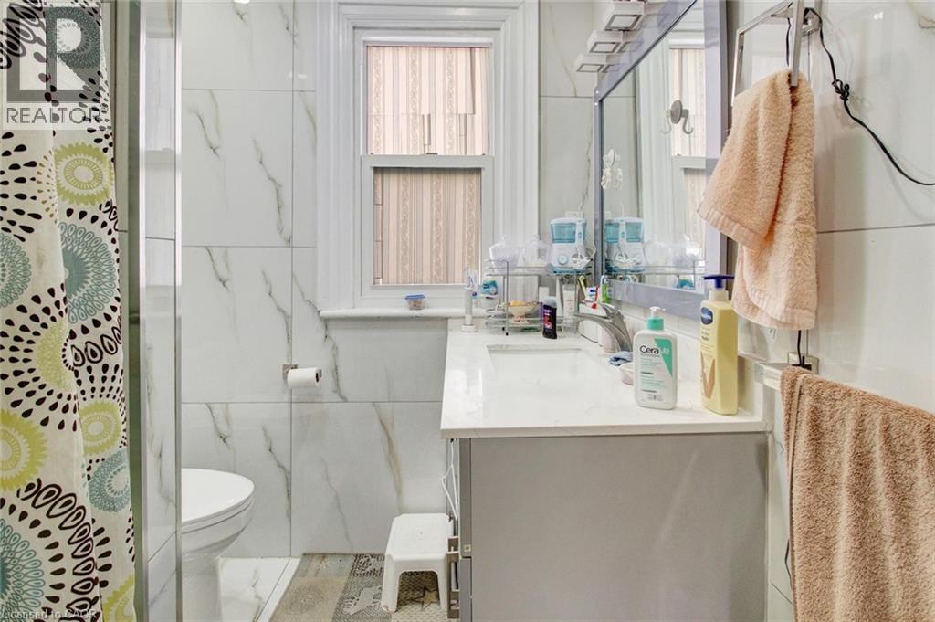 Bathroom featuring a vanity with an integrated sink, a rectangular mirror with integrated lighting, tiled walls, wood-finish flooring, and a single window - 823 Queensdale Avenue E, Hamilton, ON - Indoor Photo Showing Bathroom