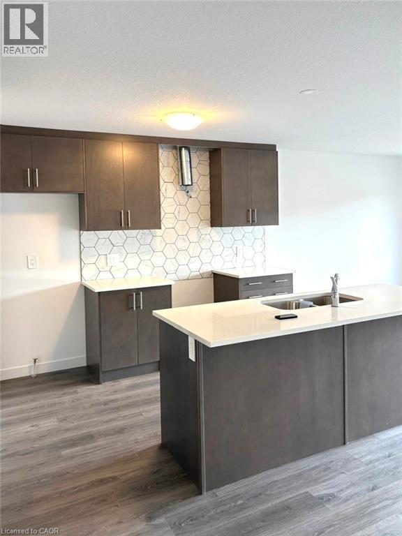 Kitchen with dark brown cabinetry, light stone counters, backsplash, dark wood finished floors, and a center island with sink - 7 Pin Oak Common, Cambridge, ON - Indoor Photo Showing Kitchen With Double Sink