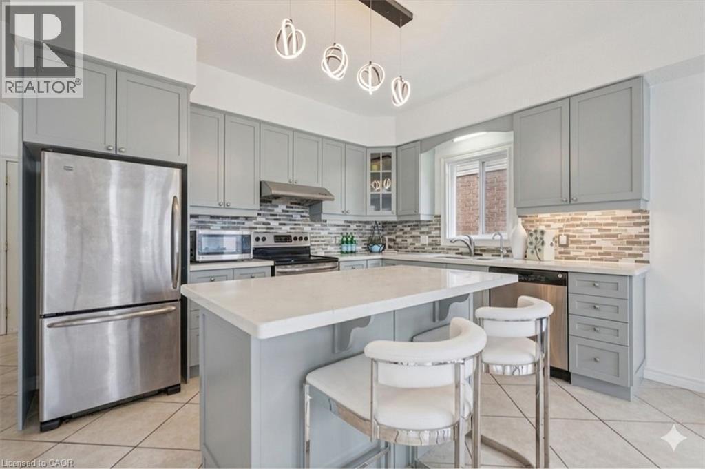 Contemporary kitchen featuring a central island with a light-toned countertop, light grey cabinetry, tile flooring, stainless steel appliances, and a mosaic tile backsplash - 512 Wood Nettle Way, Waterloo, ON - Indoor Photo Showing Kitchen With Upgraded Kitchen