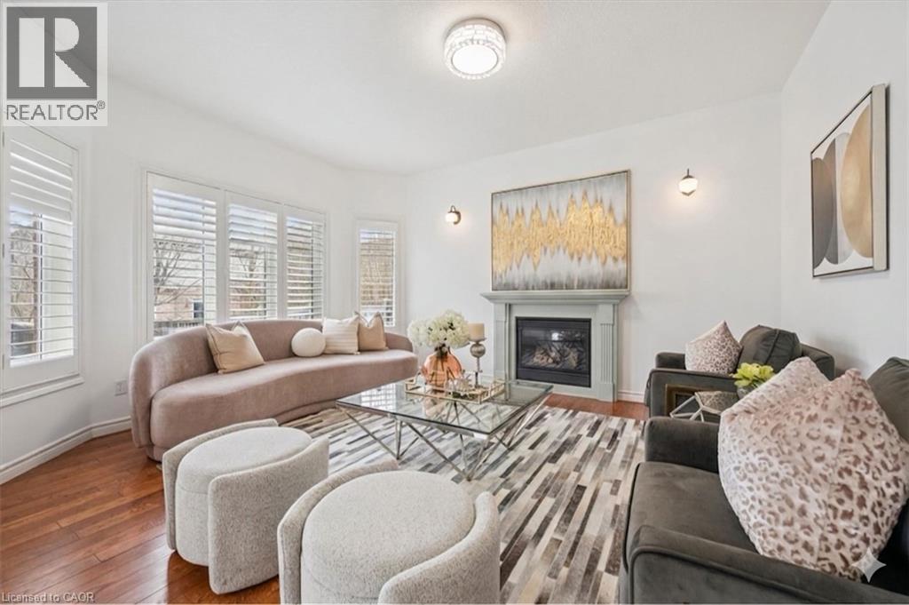 Living area featuring wood-finish flooring, a fireplace with a light-colored mantel, wall sconce lighting, white plantation shutters, and a modern ceiling fixture - 512 Wood Nettle Way, Waterloo, ON - Indoor With Fireplace