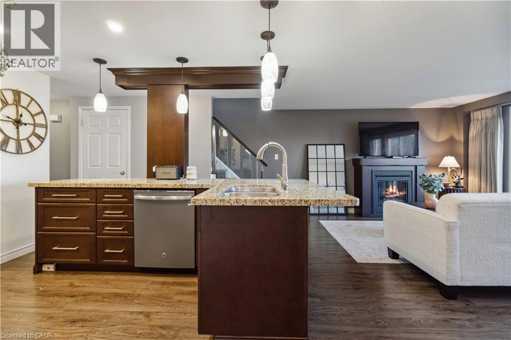 Kitchen featuring an island with sink, dark wood finish cabinets, stainless steel dishwasher, open floor plan, and a glass covered fireplace - 27 Red Clover Crescent, Kitchener, ON - Indoor With Fireplace