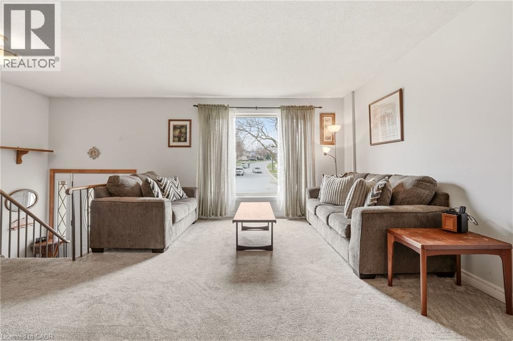 Living room featuring light-colored carpet flooring, a large window with drapes, and a partial view of a staircase with a metal railing - 2349 Middlesmoor Crescent, Burlington, ON - Indoor Photo Showing Other Room