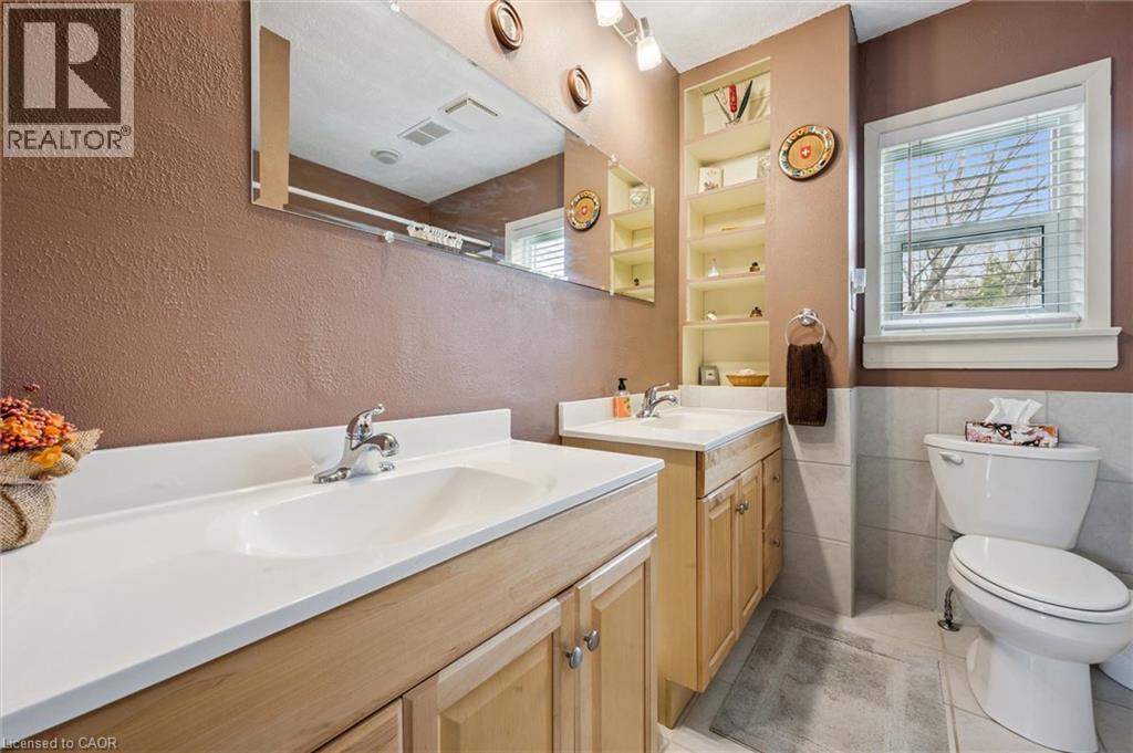 This bathroom features dual vanity sinks with light wood cabinetry, a large mirror, built-in shelving, and a window providing natural light - 155 Hungerford Road, Cambridge, ON - Indoor Photo Showing Bathroom
