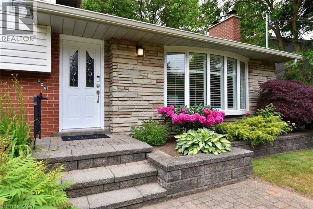 The property features a brick and stone facade with a white front door, a bay window with blinds, and mature landscaping - 626 Vanderburgh Drive, Burlington, ON - Outdoor