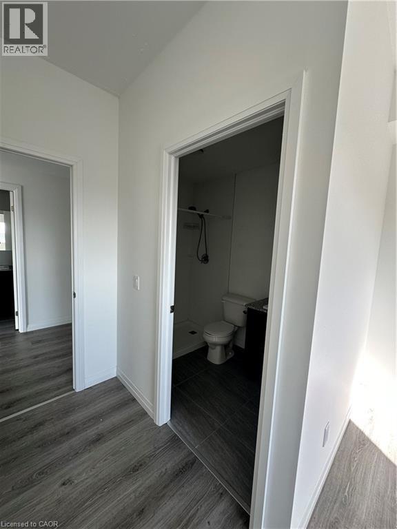 The property features light-colored walls and dark wood-look flooring throughout, with a bathroom visible through a doorway showcasing dark tiled flooring, a toilet, and a shower area - 108 Garment Street S Unit# 708, Kitchener, ON - Indoor Photo Showing Other Room