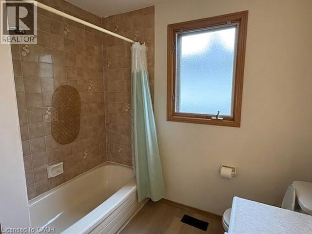 Bathroom with a shower/tub combination featuring tan tile surround, a frosted glass window with wood trim, light beige walls, wood-finish flooring, and a toilet with a light-colored countertop - 70 Bechtel Drive, Kitchener, ON - Indoor Photo Showing Bathroom