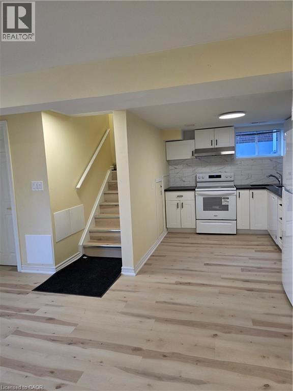 The property features light-toned flooring, a staircase with a handrail, and a kitchen area with white cabinetry, a white stove, and a tiled backsplash - 62 Richwill Road Unit# Lower, Hamilton, ON - Indoor
