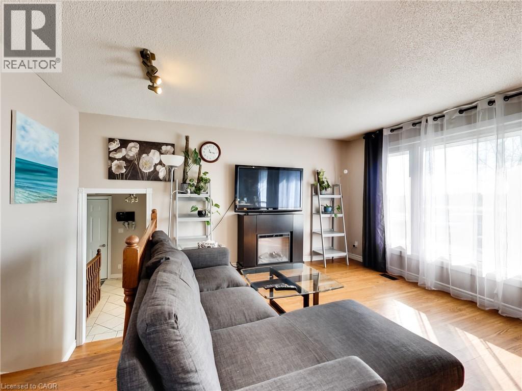 Living area featuring wood-finish flooring, a wall-mounted fireplace with a dark surround, and large windows with sheer and blackout curtains - 9183 Silver Street, Caistor Centre, ON - Indoor Photo Showing Living Room