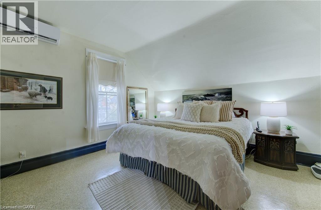 This inviting room features a window with white curtains, a wall-mounted air conditioning unit, and a bed with a decorative headboard - 48 Dupont Street, Waterloo, ON - Indoor Photo Showing Bedroom