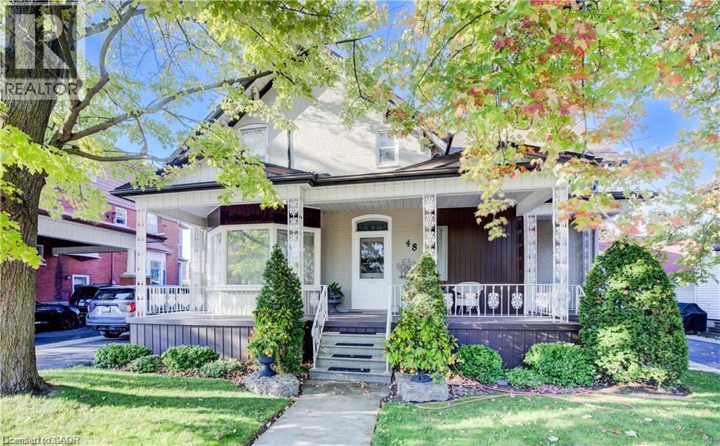 The property features a charming front porch with decorative white railings and columns, a bay window, and a neatly manicured lawn with mature trees - 48 Dupont Street, Waterloo, ON - Outdoor With Deck Patio Veranda