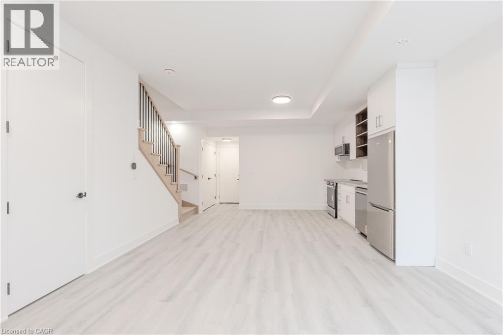 Open concept living space featuring light-toned flooring, a light wood staircase with dark balusters, and a compact kitchen area equipped with stainless steel appliances and white cabinetry - 144 Everett Common, St. Catharines, ON - Indoor