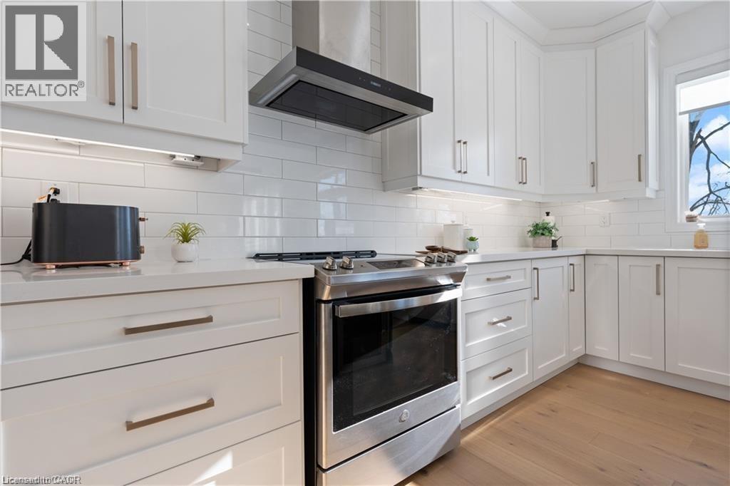 Kitchen with stainless steel gas range, white cabinetry, light wood-type flooring, backsplash, and light stone countertops - 66 Cesar Place, Ancaster, ON - Indoor Photo Showing Kitchen With Upgraded Kitchen