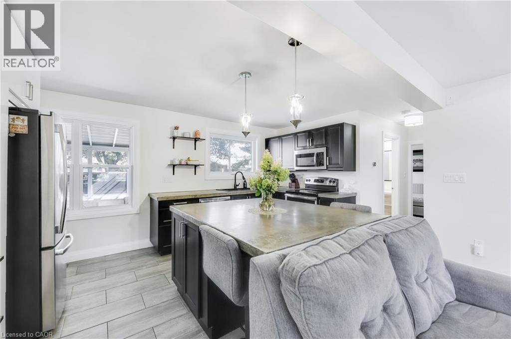 Open-concept kitchen featuring dark cabinetry, stainless steel appliances, a central island with seating, pendant lighting, and light-toned tile flooring - 18 Tweedsmuir Avenue, London, ON - Indoor Photo Showing Kitchen