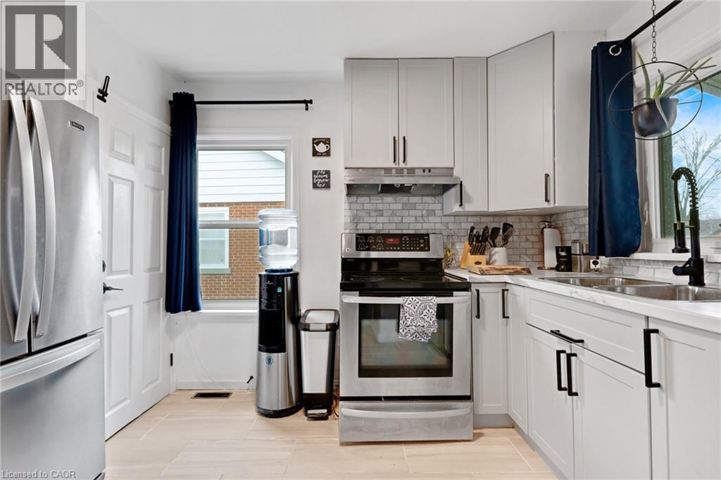 Modern kitchen featuring light-toned cabinetry with black hardware, stainless steel appliances, a subway tile backsplash, a double basin sink with a black coiled faucet, and wood-finish flooring - 34 Christopher Drive, Cambridge, ON - Indoor Photo Showing Kitchen With Double Sink