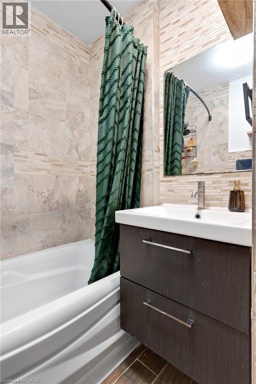 Bathroom featuring a white bathtub, full-height wall tiling, a rectangular mirror, a wall-mounted vanity with two drawers, and wood-finish flooring - 34 Christopher Drive, Cambridge, ON - Indoor Photo Showing Bathroom