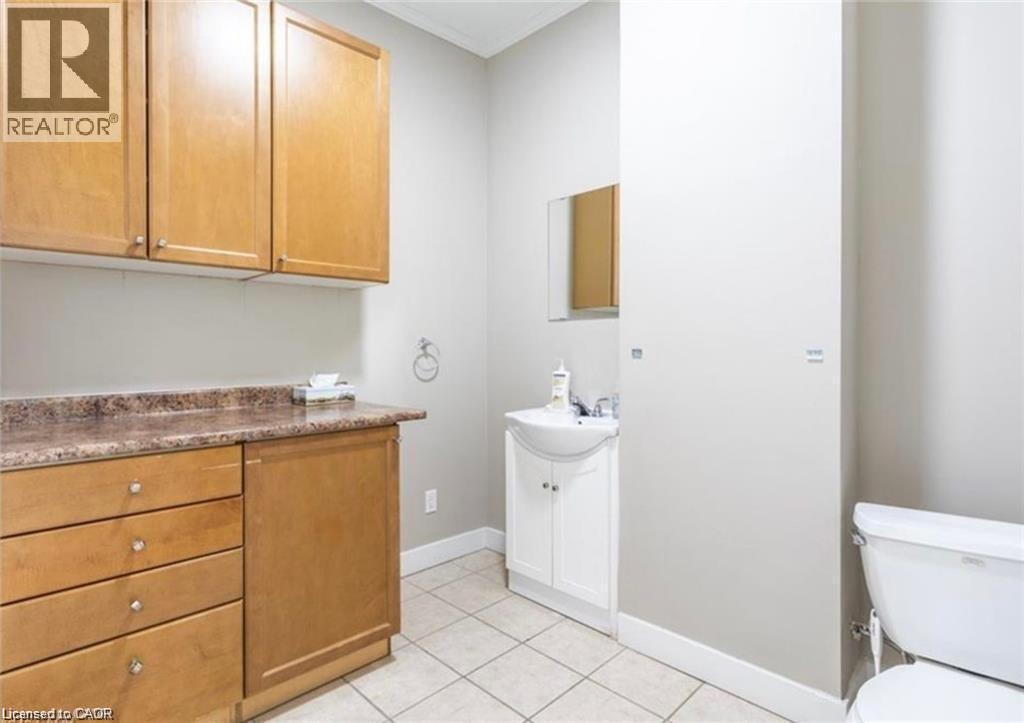 This room features light-colored floor tiles, a toilet, a vanity with a white basin and white cabinets, and wooden cabinetry with a countertop - 833 King Street E, Hamilton, ON