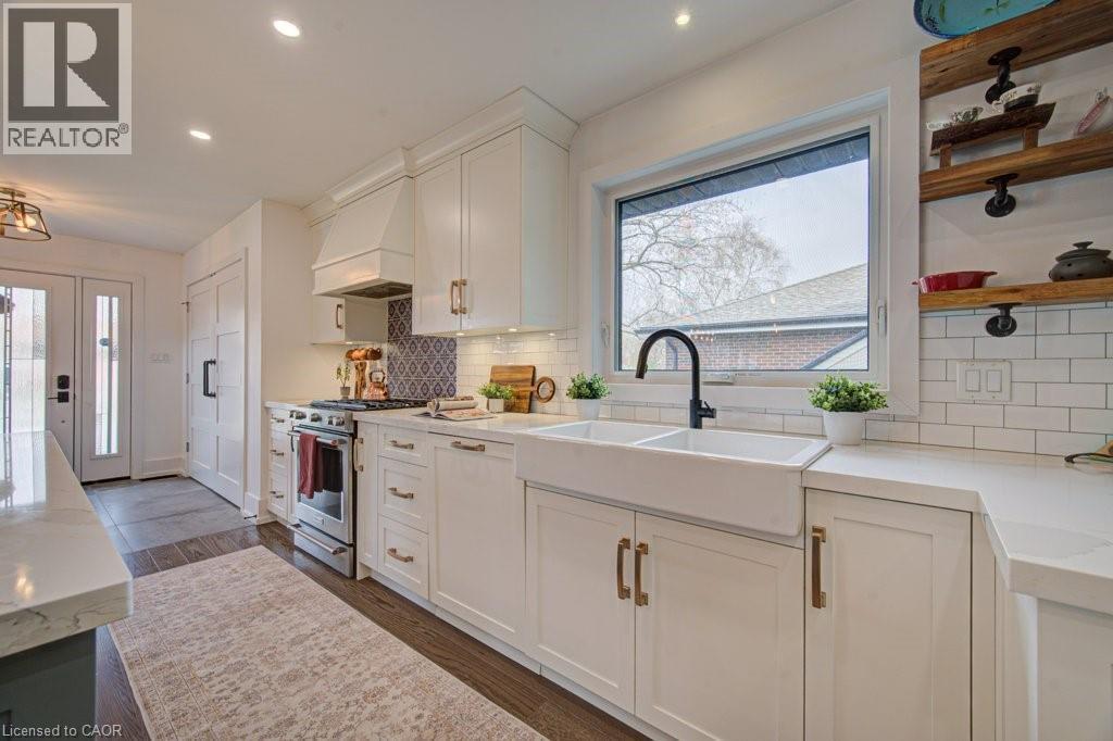 Contemporary kitchen featuring white shaker cabinetry, an integrated range hood, stainless steel appliances, a double basin farmhouse sink with a matte black faucet, and open wood shelving - 1486 Augustine Drive, Burlington, ON - Indoor Photo Showing Kitchen With Double Sink With Upgraded Kitchen