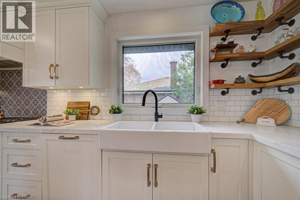 Kitchen featuring a double basin farmhouse sink with a matte black faucet, light-toned countertops, white shaker cabinetry with gold-tone hardware, white subway tile backsplash, and wood-finish open s - 1486 Augustine Drive, Burlington, ON - Indoor Photo Showing Kitchen With Double Sink