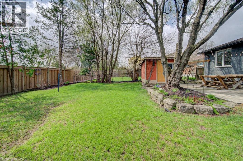 Expansive lawn area featuring a wooden privacy fence, mature trees, a stone-lined garden bed, a brightly colored shed, and a concrete patio - 1486 Augustine Drive, Burlington, ON - Outdoor