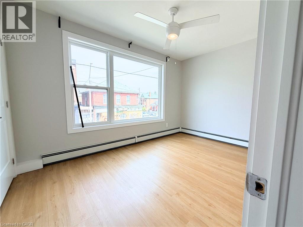 Bright room featuring a wide window, wood-finish flooring, a ceiling fan with integrated lighting, and baseboard heating - 310 Barton Street E Unit# A, Hamilton, ON - Indoor Photo Showing Other Room