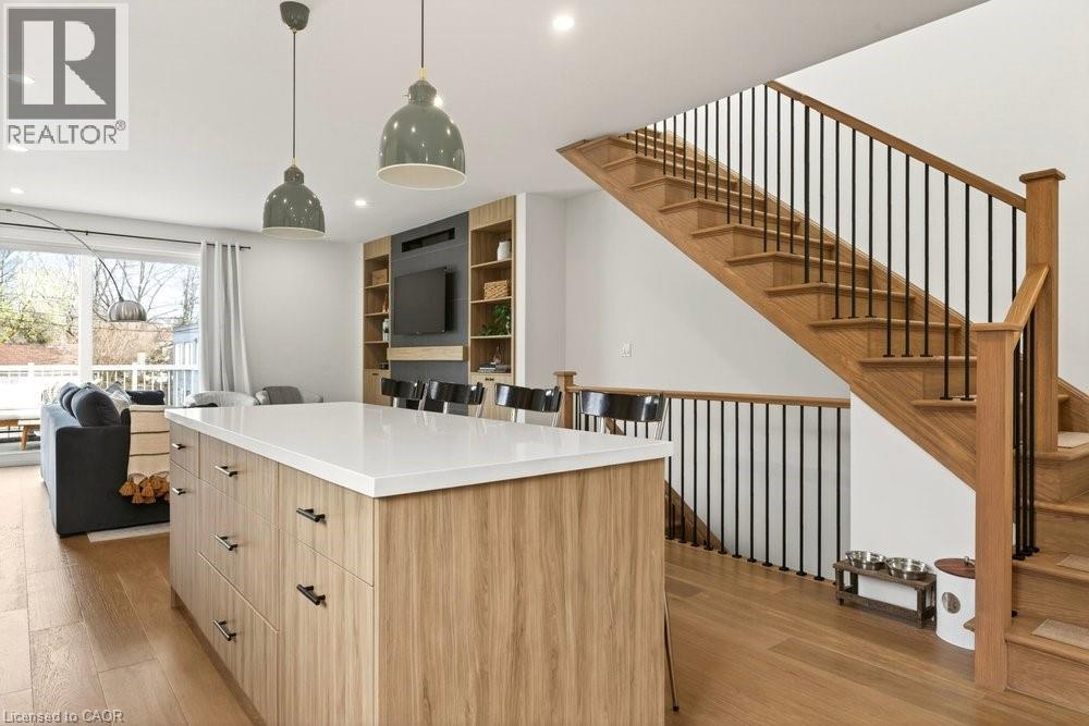 Open-concept living space featuring wood-finish flooring, a contemporary kitchen island with a light-toned countertop, a built-in media center with shelving, and a prominent staircase with wood treads - 128 Canada Street, Hamilton, ON - Indoor