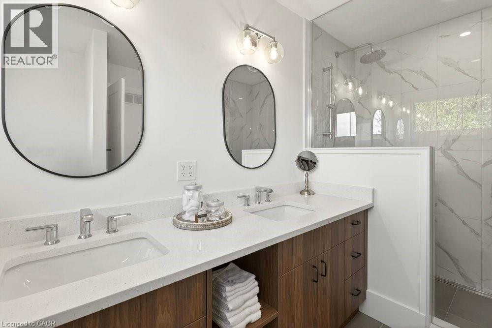 Bathroom featuring a dual-sink wood-finish vanity with stone-look countertops, chrome faucets, and two oval mirrors - 128 Canada Street, Hamilton, ON - Indoor Photo Showing Bathroom
