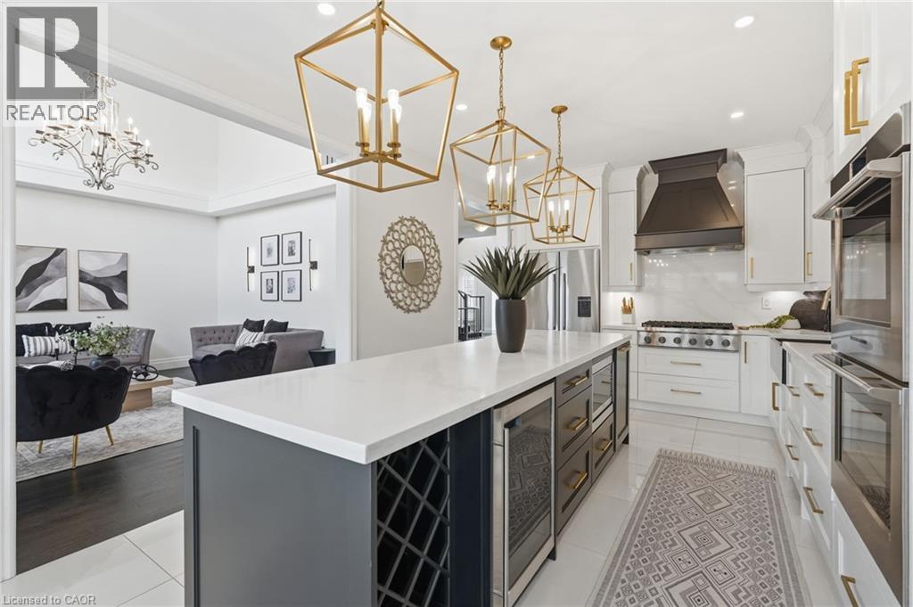 Gourmet kitchen featuring a substantial island with integrated wine storage and beverage coolers, pristine white countertops, white cabinetry with gold-tone hardware, and a prominent range hood over a - 47 Winners Way, Binbrook, ON - Indoor Photo Showing Kitchen With Upgraded Kitchen