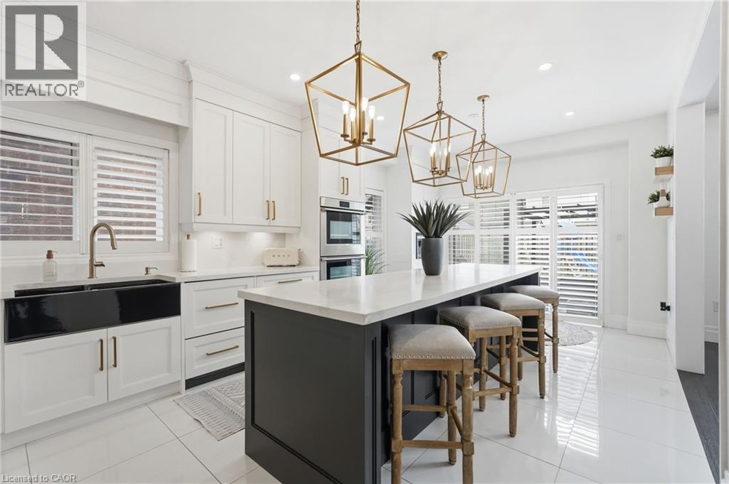 Spacious kitchen featuring an extended island with seating, white shaker cabinetry, stainless steel double wall ovens, a large black farmhouse sink, and polished white tile flooring - 47 Winners Way, Binbrook, ON - Indoor
