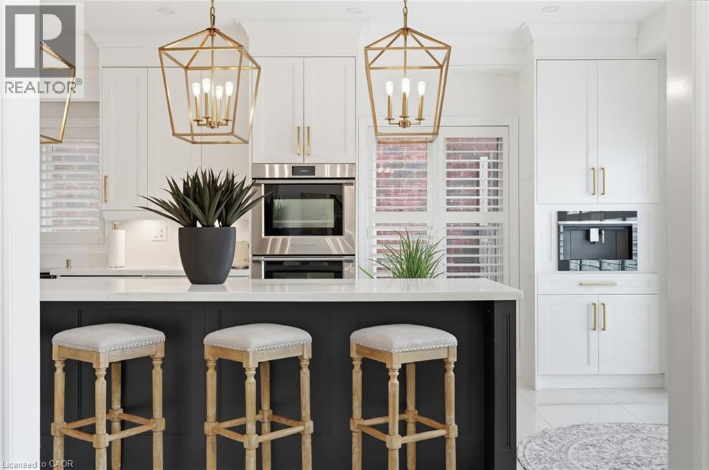Kitchen featuring a dark-toned island with a light-toned countertop, white cabinetry with gold-toned hardware, built-in stainless steel oven, and two geometric gold-toned pendant lights - 47 Winners Way, Binbrook, ON - Indoor