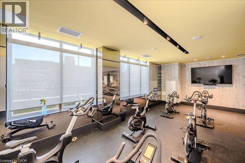 Fitness studio featuring multiple stationary bicycles, expansive windows with roller shades, a large wall-mounted display, wood-finish wall panels, and recessed lighting - 85 Duke Street W Unit# 507, Kitchener, ON - Indoor Photo Showing Gym Room
