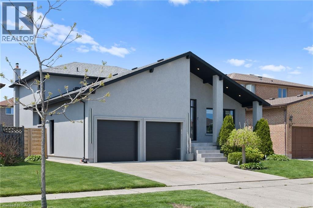 Modern stucco exterior with a prominent A-frame entry, featuring a two-car garage with dark doors, a concrete driveway, and landscaped front yard with mature shrubbery and a young tree - 17 Clearview Drive, Hamilton, ON - Outdoor