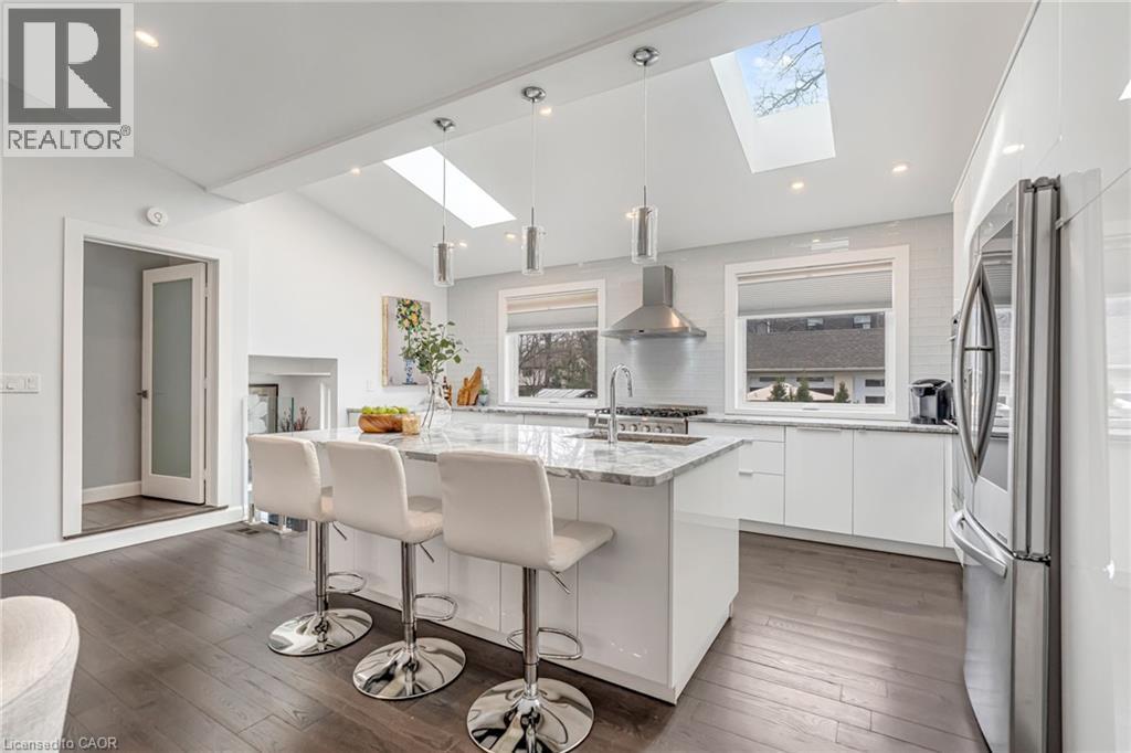 Open-concept kitchen featuring an oversized island with seating, a vaulted ceiling with skylights, stainless steel appliances, and wood-finish flooring - 2123 Deyncourt Drive, Burlington, ON - Indoor