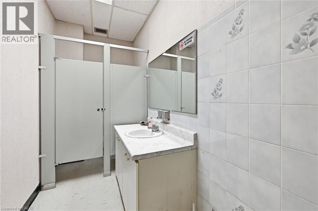 Restroom featuring white privacy partitions, a vanity with an integrated sink, a wall-mounted mirror, and white tiled walls with subtle floral accents - 869 Barton Street E, Hamilton, ON
