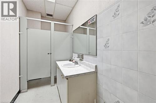 Restroom featuring white privacy partitions, a vanity with an integrated sink, a wall-mounted mirror, and white tiled walls with subtle floral accents - 869 Barton Street E, Hamilton, ON 