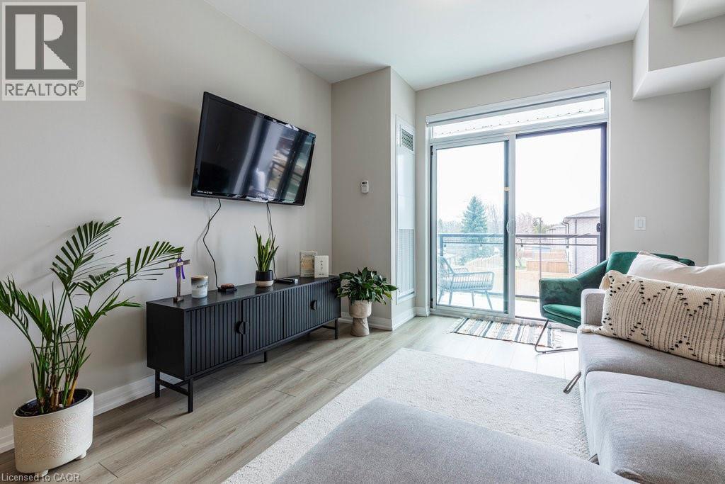 Bright living area featuring wood-finish flooring, a contemporary console cabinet, and a sliding glass door leading to a private balcony with black metal railings - 120 Springvalley Crescent Unit# 208, Hamilton, ON - Indoor