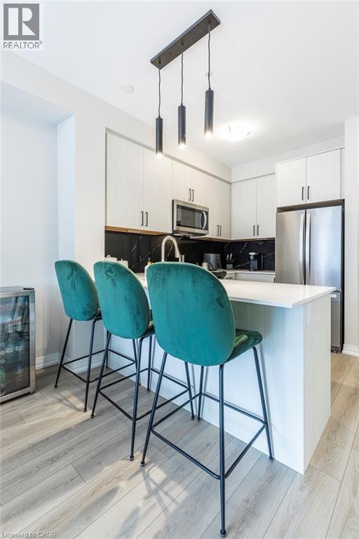 Modern kitchen featuring an island with white countertop, contemporary pendant lighting, light wood-finish flooring, white cabinetry, and stainless steel appliances - 120 Springvalley Crescent Unit# 208, Hamilton, ON - Indoor Photo Showing Kitchen With Stainless Steel Kitchen With Upgraded Kitchen