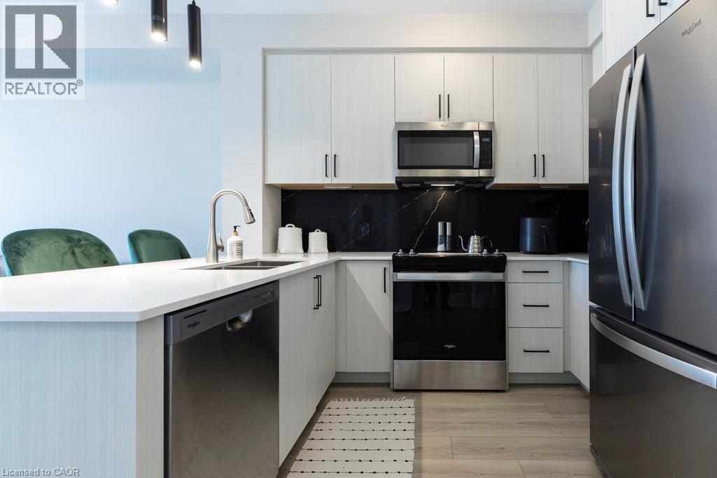Modern kitchen featuring light wood-finish cabinetry, stainless steel appliances, white countertops, and a black backsplash with subtle veining - 120 Springvalley Crescent Unit# 208, Hamilton, ON - Indoor Photo Showing Kitchen With Stainless Steel Kitchen With Upgraded Kitchen