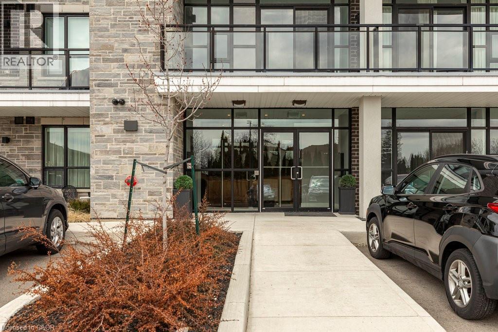 Modern building facade featuring a stone veneer exterior, expansive glass entrance doors, black-framed windows, and balconies with black railings - 120 Springvalley Crescent Unit# 208, Hamilton, ON - Outdoor With Balcony