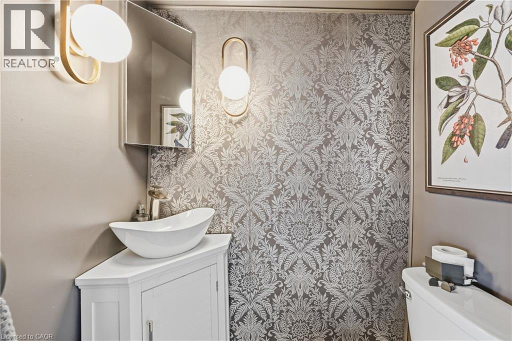 Powder room featuring striking patterned wallpaper, a white vessel sink atop a corner vanity, a wall-mounted mirror, and contemporary sconce lighting - 361 Cumberland Avenue, Hamilton, ON - Indoor Photo Showing Bathroom