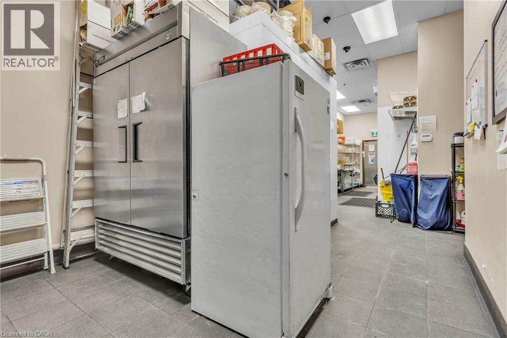 Commercial kitchen area featuring a large, stainless steel double-door refrigerator, a white upright refrigerator, and grey tile flooring - 589 Fairway Road S, Kitchener, ON