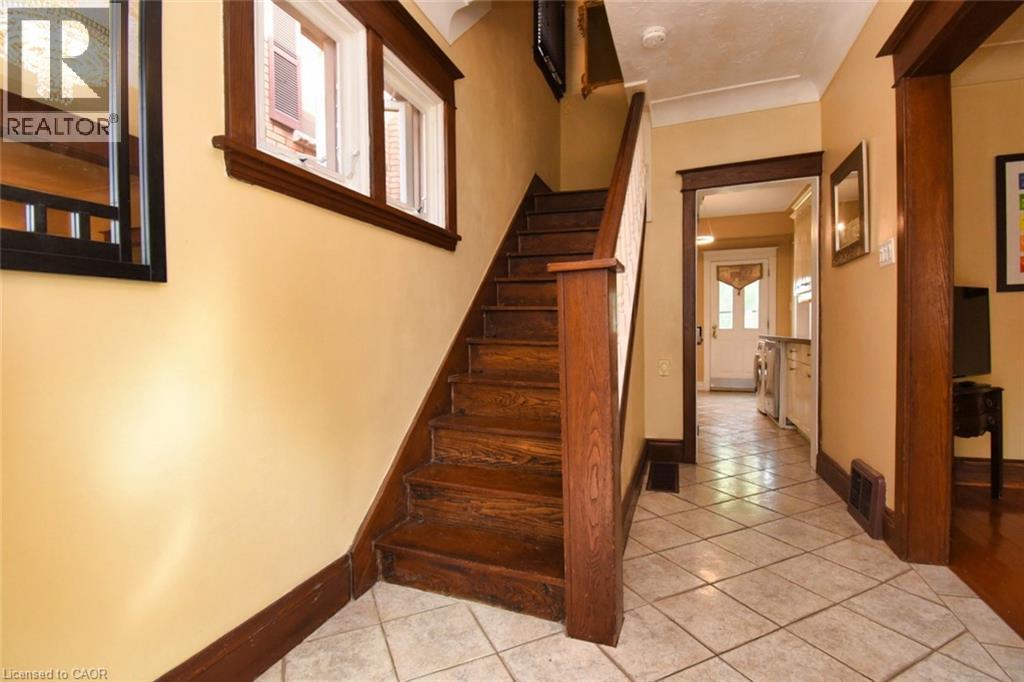 Wood staircase with a contrasting white baluster, tile flooring, and a framed mirror reflecting a chandelier - 522 Aberdeen Avenue, Hamilton, ON - Indoor Photo Showing Other Room