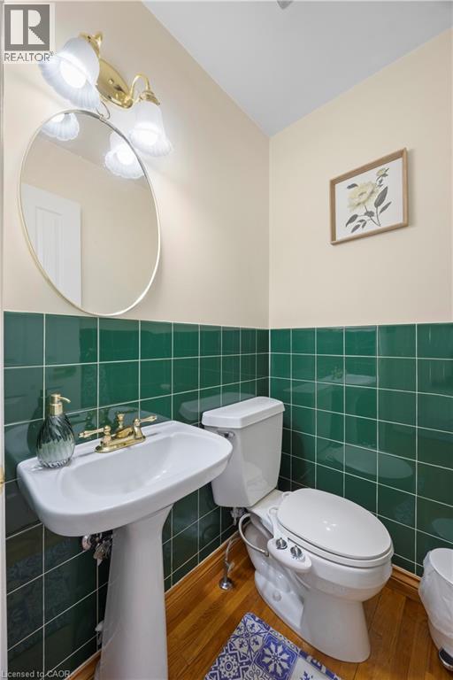 Pedestal sink with brass-finish faucet, green tiled wainscoting, circular wall mirror, and wood-finish flooring - 277 Blackacres Boulevard, London, ON - Indoor Photo Showing Bathroom